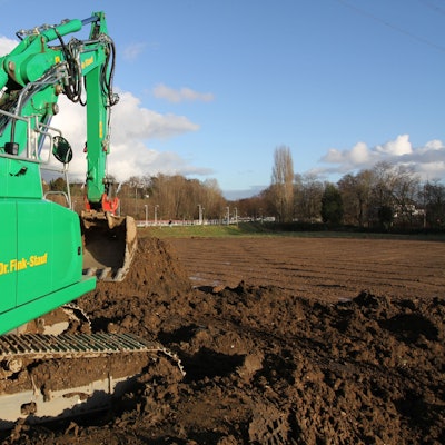 Ein grüner Bagger vor einem unbebautem Feld.