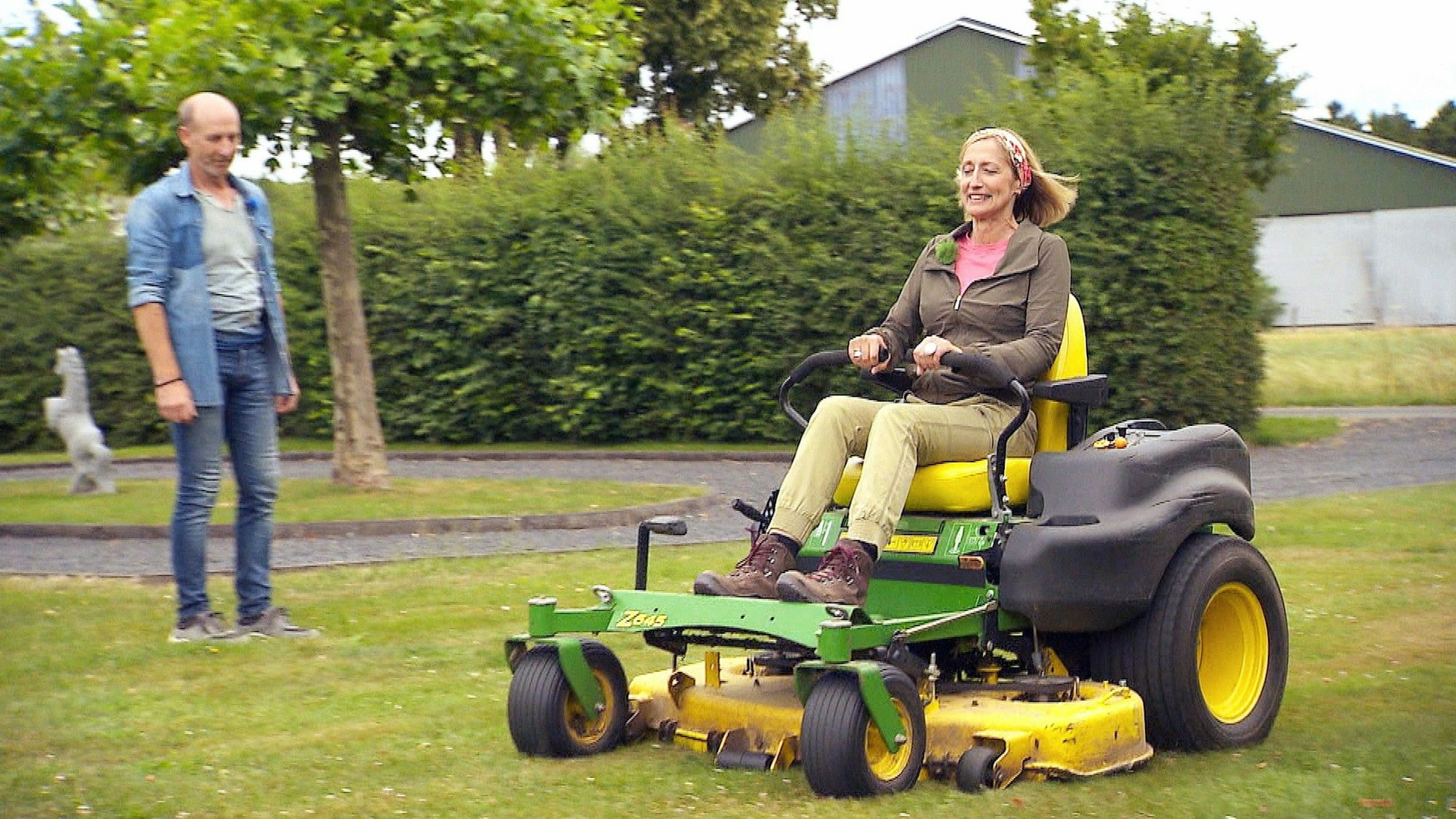 Eine Frau bedient einen Aufsitzrasenmäher, ein Mann schaut ihr dabei zu.