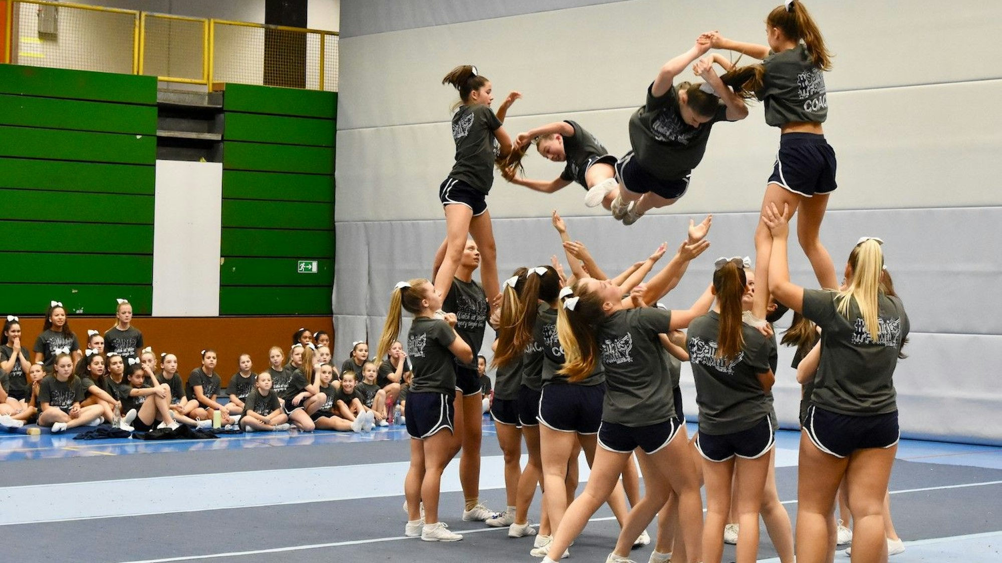 Eine Gruppe junger Frauen zeigt eine Choreographie in ihrem Wettkampfsport Cheerleading. Jüngere Mädchen in den gleichen Vereinstrikots schauen zu.