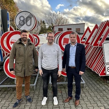 Drei Männer stehen vor einer großen Zahl aufgereihter Verkehrsschilder.