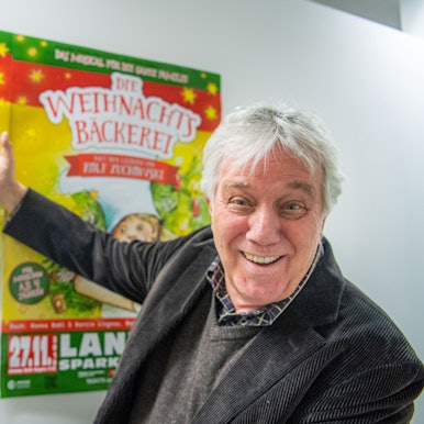 Rolf Zuckowski, Liedermacher, zeigt auf Poster seines Musicals "Die Weihnachtsbäckerei"