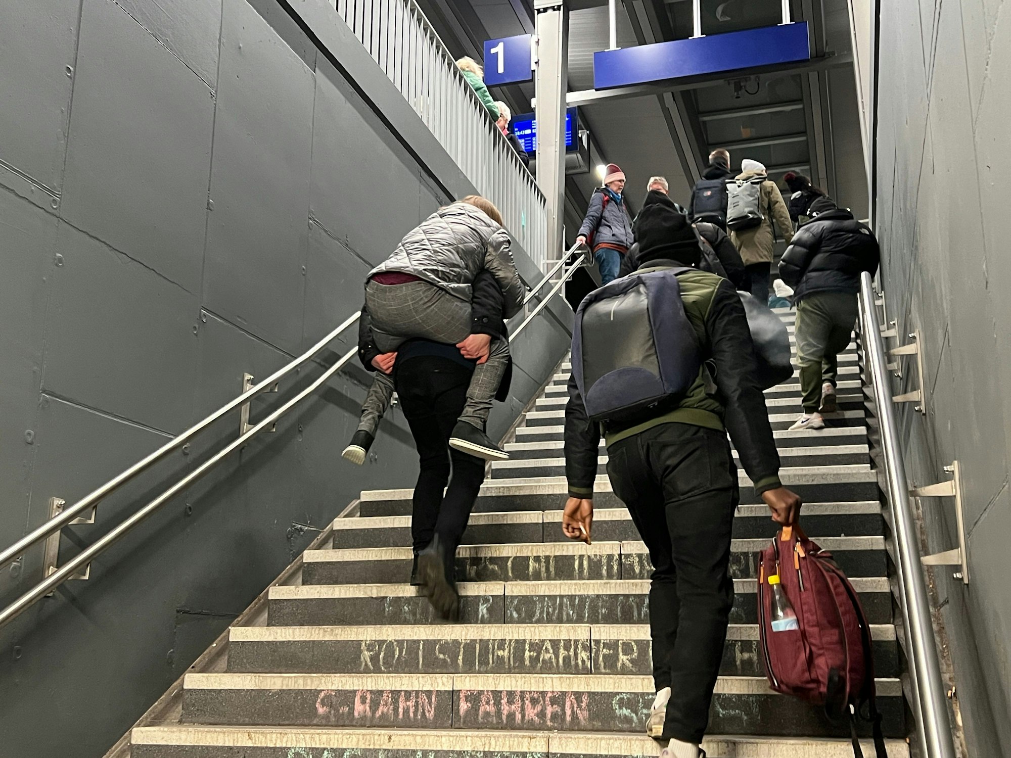 Ein Mann trägt eine gehbehinderte Frau die Treppe zum Bahnsteig hinauf.