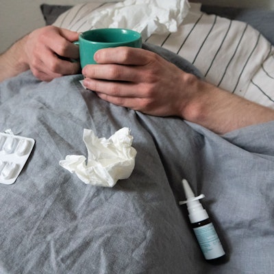 Ein Mann liegt mit Taschentüchern, Teetasse, Nasenspray und Tabletten im Bett.
