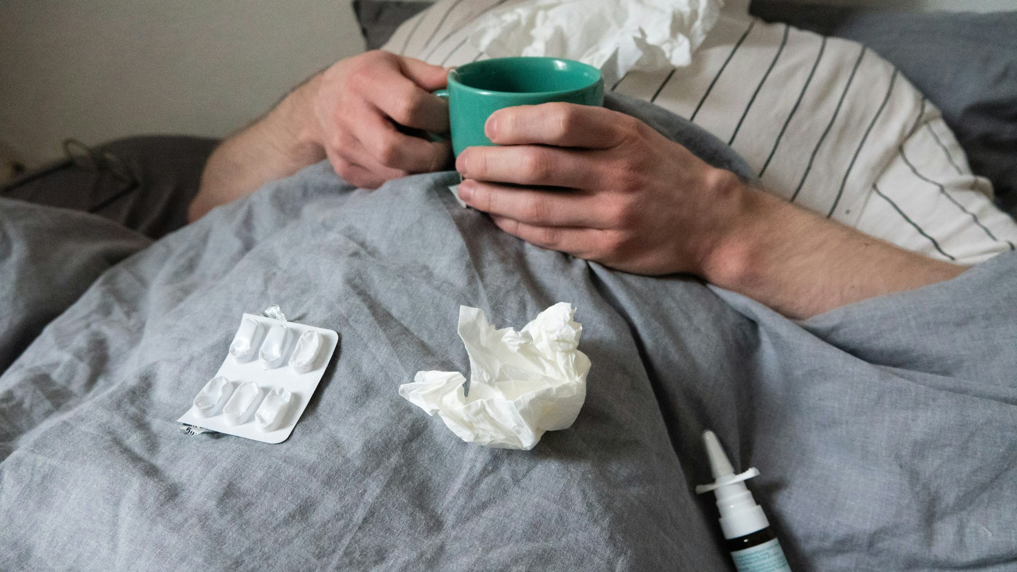 Ein Mann liegt mit Taschentüchern, Teetasse, Nasenspray und Tabletten im Bett.