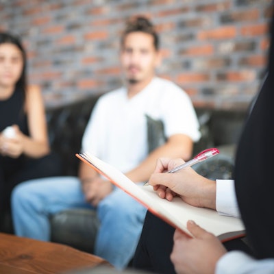 In Hintergrund unscharf zu sehen: ein Paar auf einem Sofa, das zuhört. Rechts im Vordergrund angeschnitten eine Person, die handschriftliche Aufzeichnungen anfertigt.