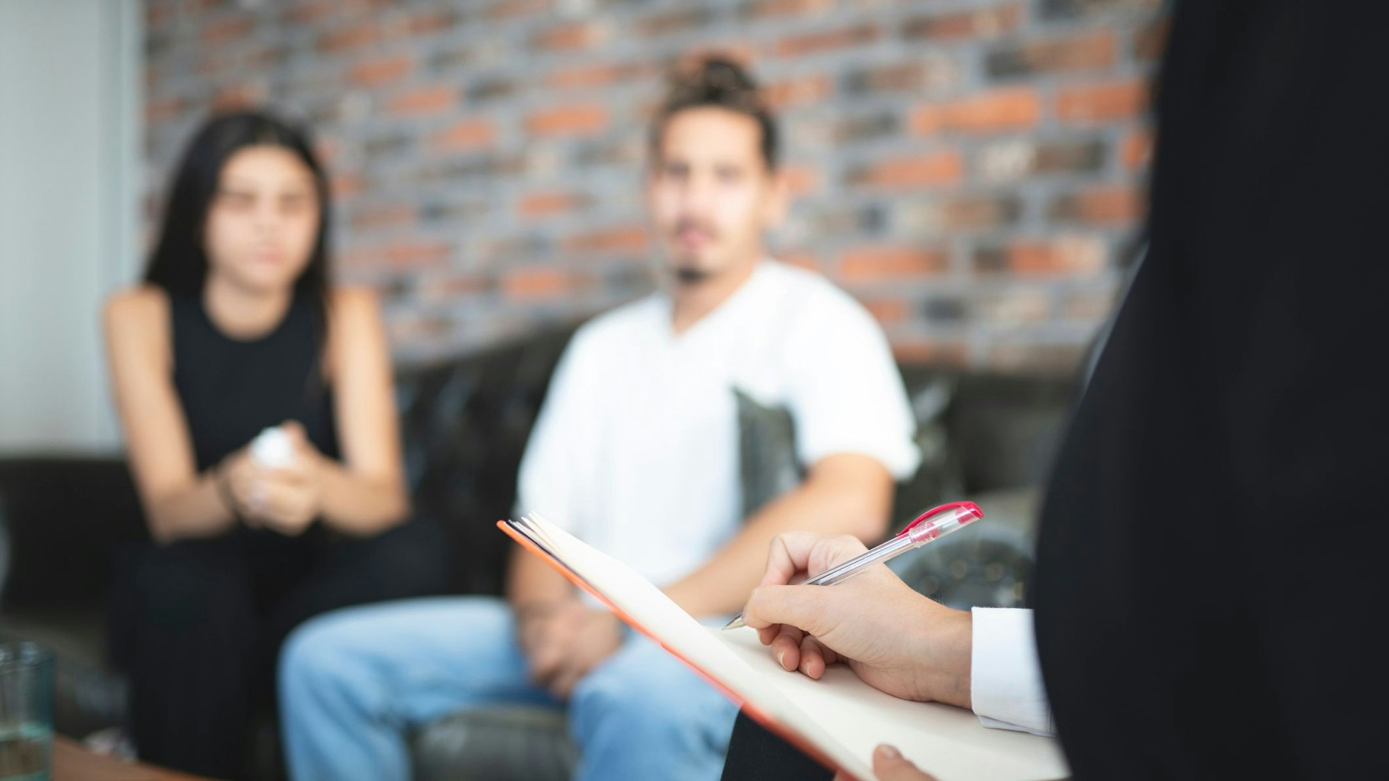 In Hintergrund unscharf zu sehen: ein Paar auf einem Sofa, das zuhört. Rechts im Vordergrund angeschnitten eine Person, die handschriftliche Aufzeichnungen anfertigt.
