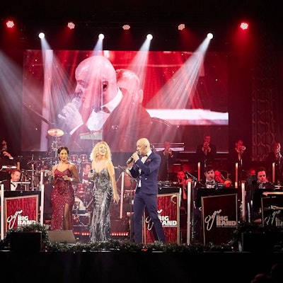 Im Vordergrund der Bühne sind Sänger Marco Matias und die Sängerinnen Sofia Andersson und Tertia Botha zu sehen, dahinter die Musiker in Uniform. Eine Großleinwand dahinter zeigt Sänger Marco Matias.