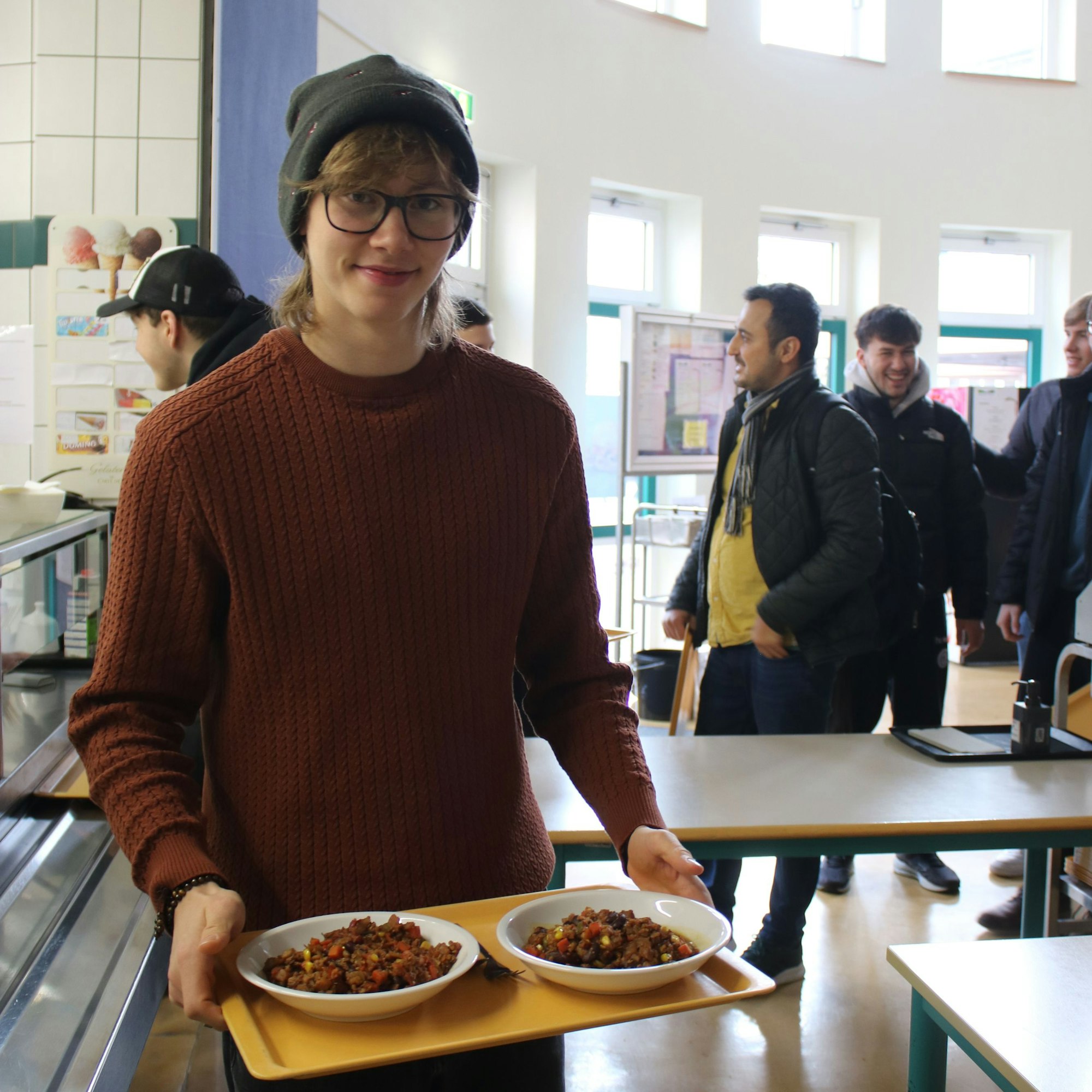 Ein junger Mann steht mit einem Tablett und Essen in einer Schulmensa.