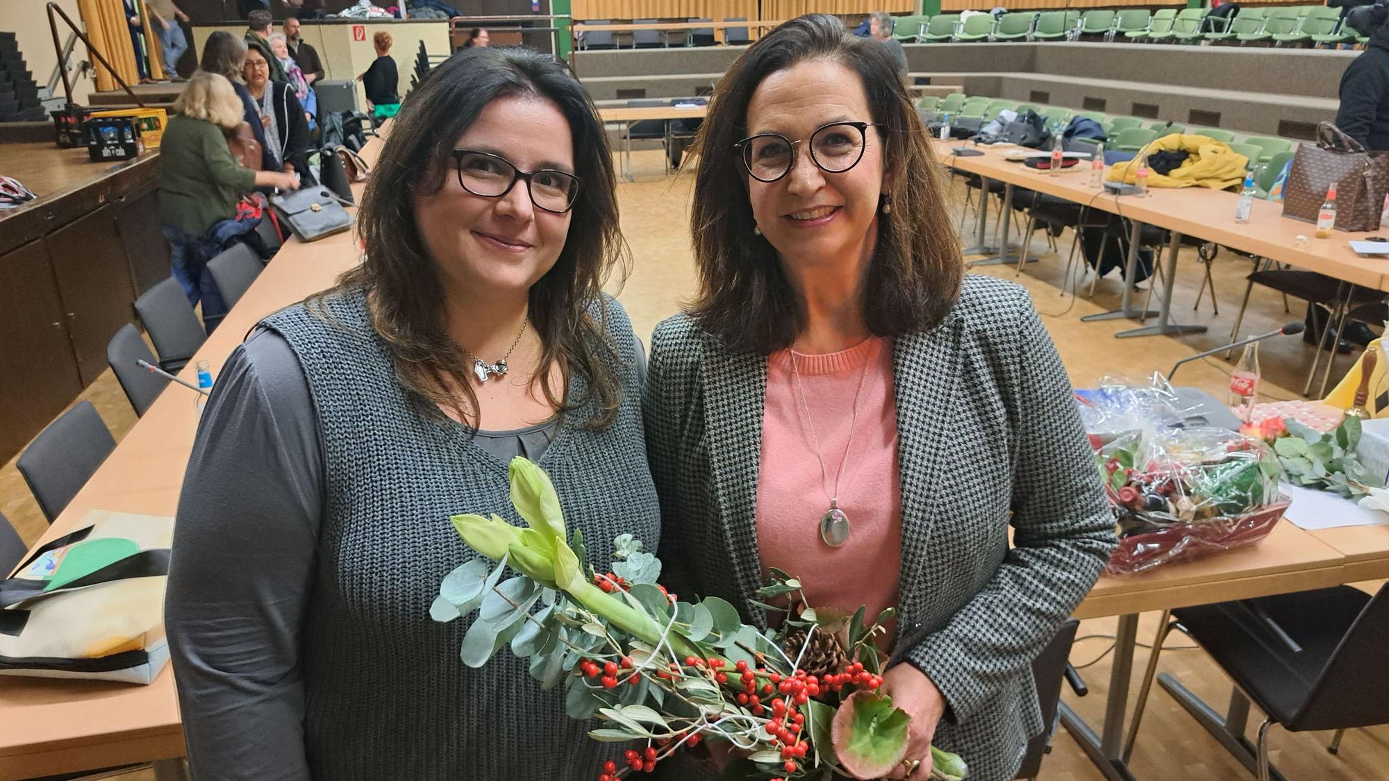 Zwei Frauen vor einem leeren Sitzungssaal. Sie lächeln; eine der beiden hat Blumen in der Hand.