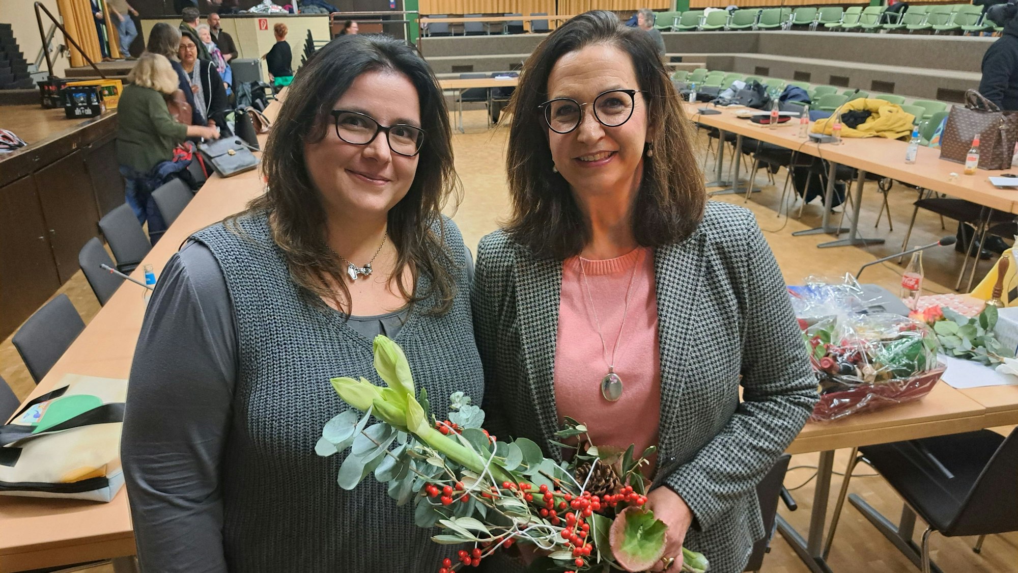 Zwei Frauen vor einem leeren Sitzungssaal. Sie lächeln; eine der beiden hat Blumen in der Hand.
