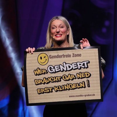 Monika Gruber steht in der Lanxess-Arena und zeigt eine Fußmatte, auf der steht: „Wer gendert, braucht gar ned erst klingeln!“