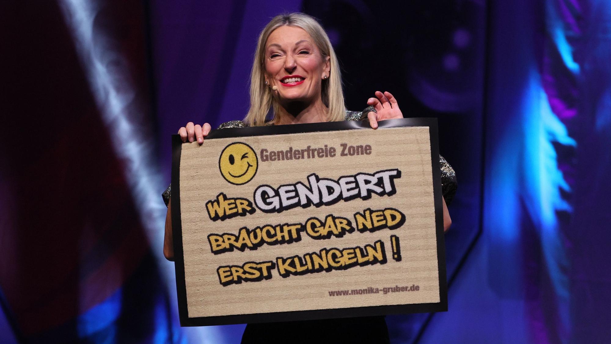 Die Kabarettistin Monika Gruber zeigt auf der Bühne eine Fußmatte, die bedruckt ist mit den Worten „Genderfreie Zone – Wer gendert, braucht gar ned erst klingeln!“