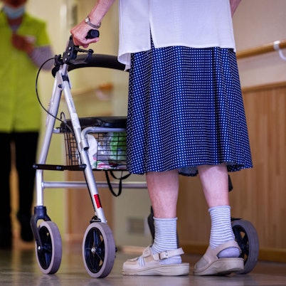 Eine Bewohnerin ist mit ihrem Rollator auf einem Flur in einem Alten- und Pflegeheim unterwegs, hinter ihr steht eine Pflegekraft.