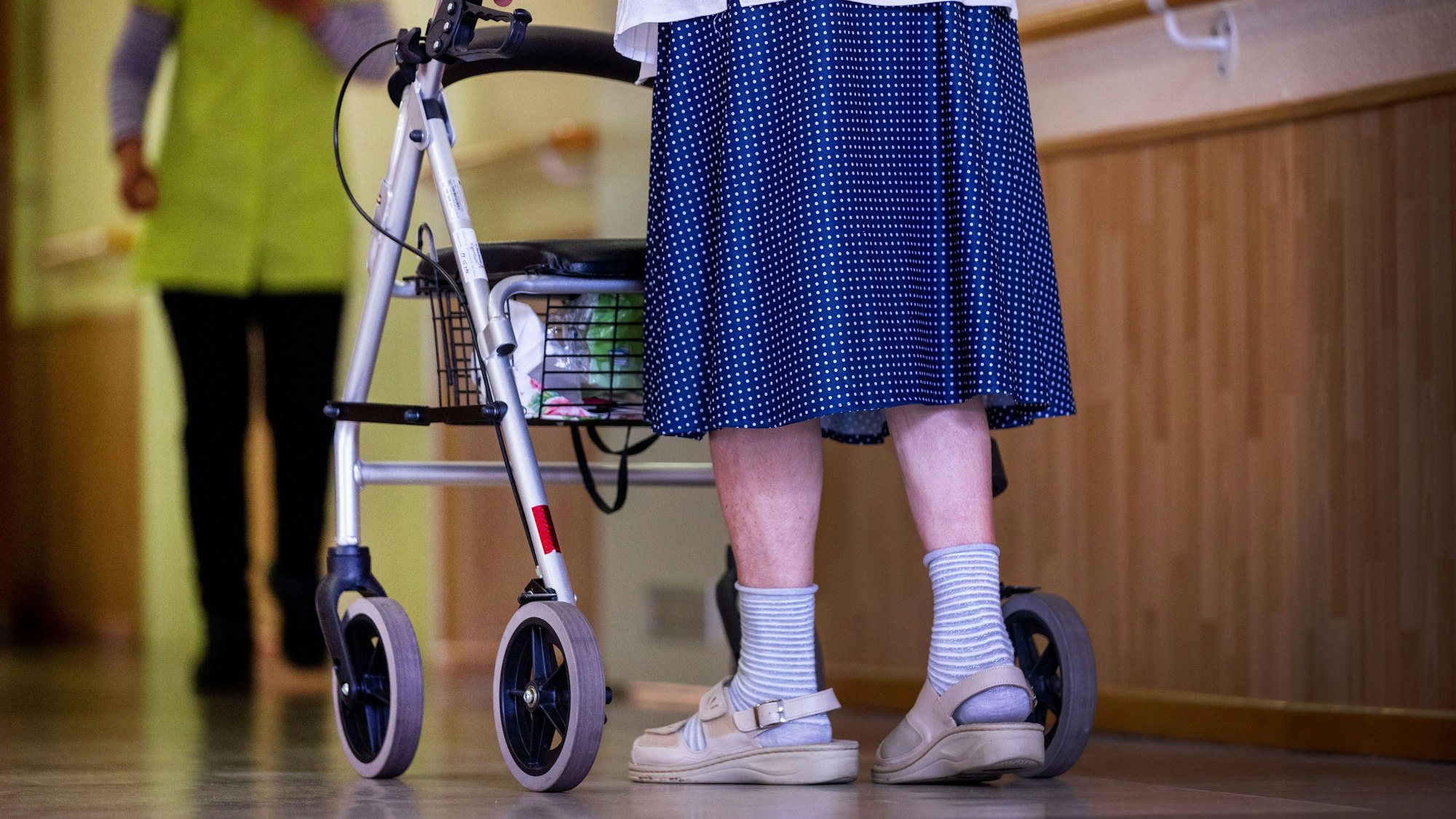 Eine Bewohnerin ist mit ihrem Rollator auf einem Flur in einem Alten- und Pflegeheim unterwegs, hinter ihr steht eine Pflegekraft.