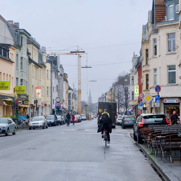 Eine Straße mit parkenden Autos am Rand und Radfahrern ist zu sehen.