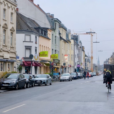 Eine Straße mit parkenden Autos am Rand und Radfahrern ist zu sehen.