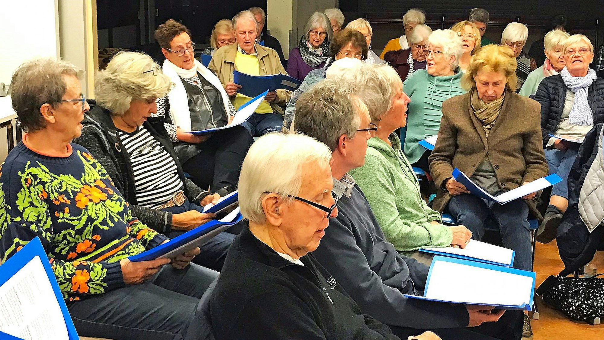 Menschen sitzen nebeneinander und singen, mit Liedmappen auf den Knien.