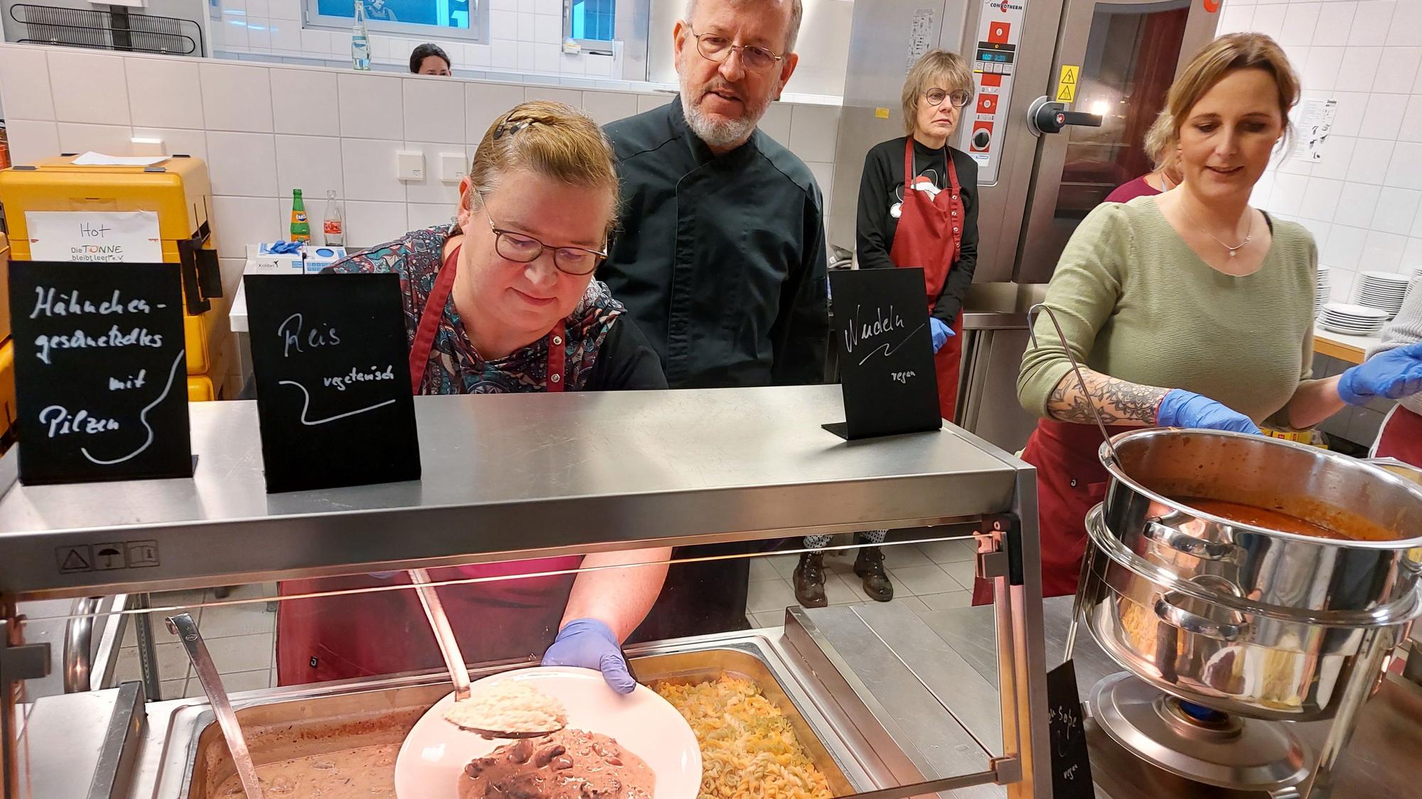 Essensausgabe in einer Mensa; eine Frau mit roter Schütze schöpft ein Essen auf einen Teller.