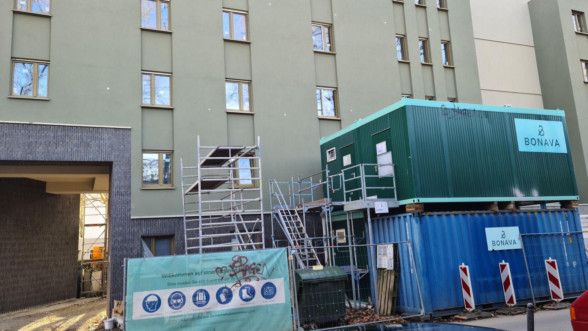 Vor einem Wohngebäude stehen ein Gerüst und Baucontainer.