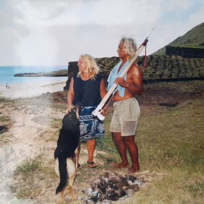 Stephanie Pauly steht mit ihrem Partner Karlo und Hund vor einer Gebetsstätte. Im Hintergrund der Strand. Karlo hat eine Harpune in der Hand.