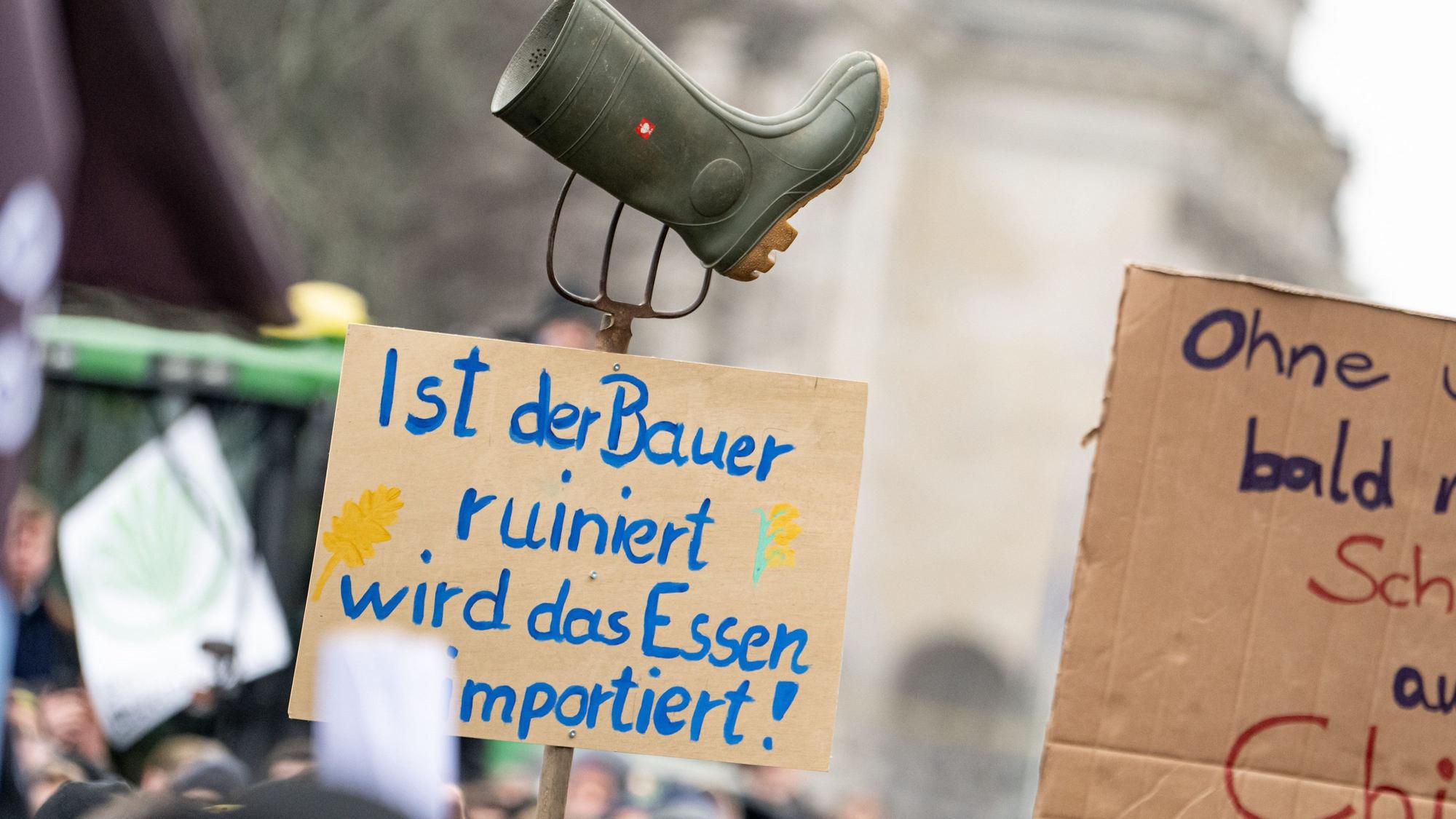 Auf einer Demonstration des Deutschen Bauernverbandes unter dem Motto „Zu viel ist zu viel! Jetzt ist Schluss!“ vor dem Brandenburger Tor in Berlin hält jemand ein Schild mit der Aufschrift „Ist der Bauer ruiniert, wird das Essen importiert“, mit eine Mistgabel und einem Gummistiefel hoch. Foto: Fabian Sommer/dpa +++ dpa-Bildfunk +++