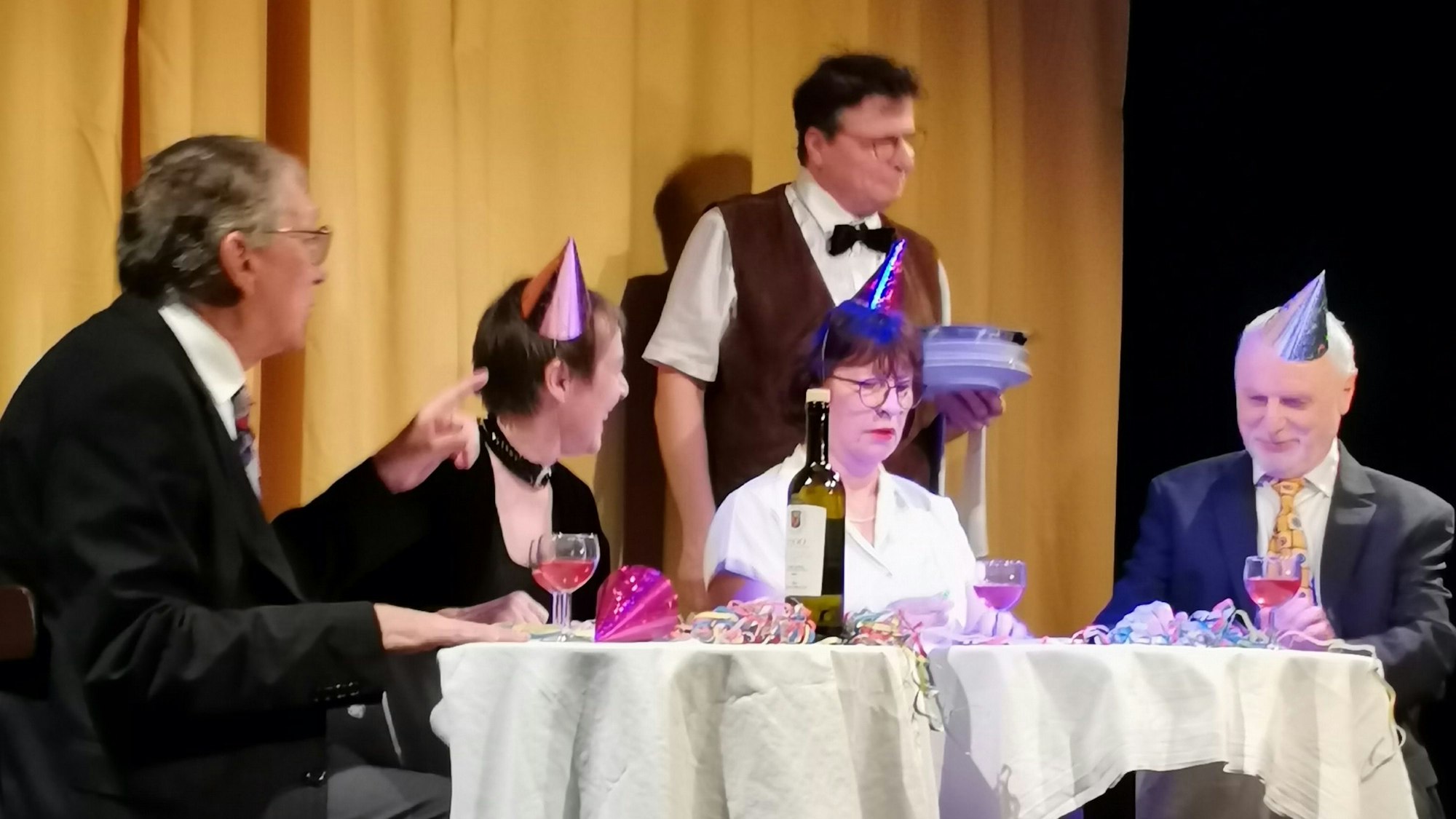 An einem Tisch sitzen zwei befreundete Ehepaare, die in einen heftigen Streit geraten. Im Hintergrund steht ein entnervt wirkender Kellner.