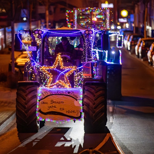 Mehre Traktoren fahren beleuchtet und mit Protestschildern versehen durch die Straßen. Auf einem steht: „Ohne Bauern geht es nicht“