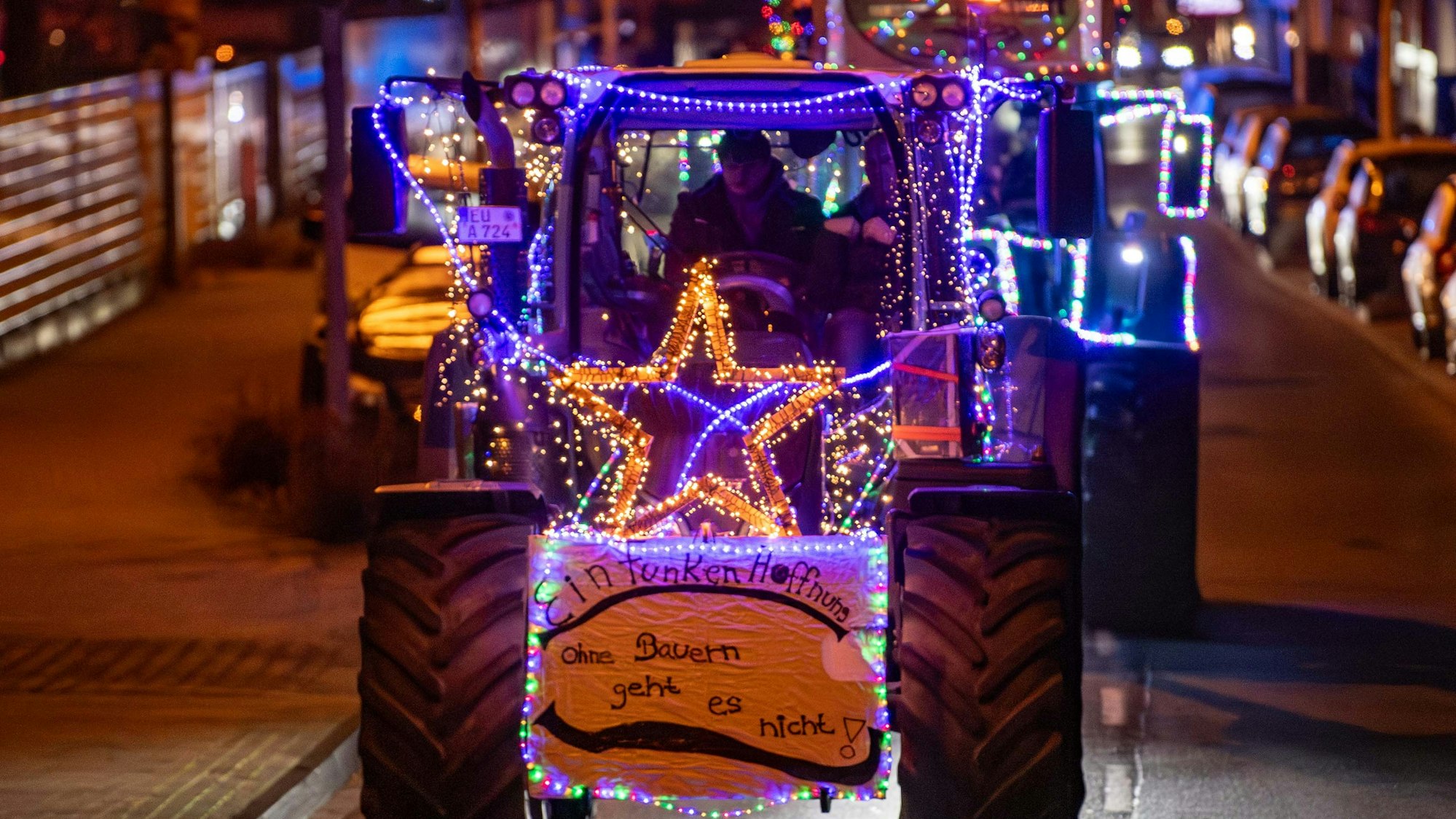 Mit Lichterketten geschmückte Traktoren fahren durch eine Straße. An einem der Fahrzeuge hängt ein Schild mit der Aufschrift „Ohne Bauern geht es nicht“.