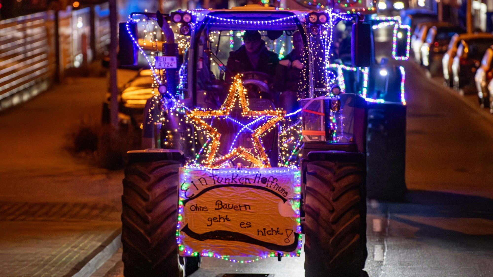 Mehre Traktoren fahren beleuchtet und mit Protestschildern versehen durch die Straßen. Auf einem steht: „Ohne Bauern geht es nicht“