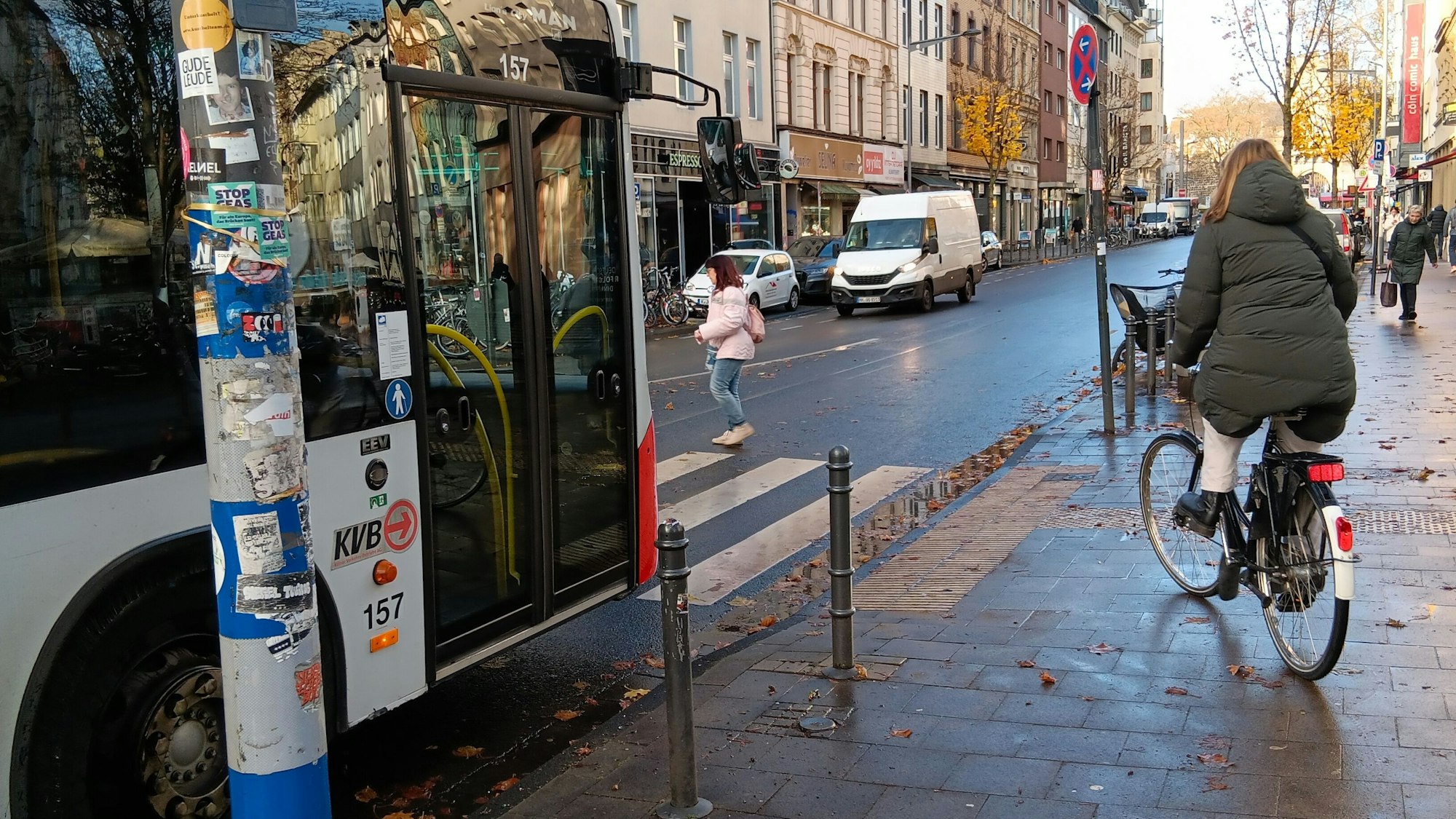 Eine Frau und eine Radfahrerin queren die Bonner Straße auf einem Zebrastreifen, vor dem ein Bus wartet.