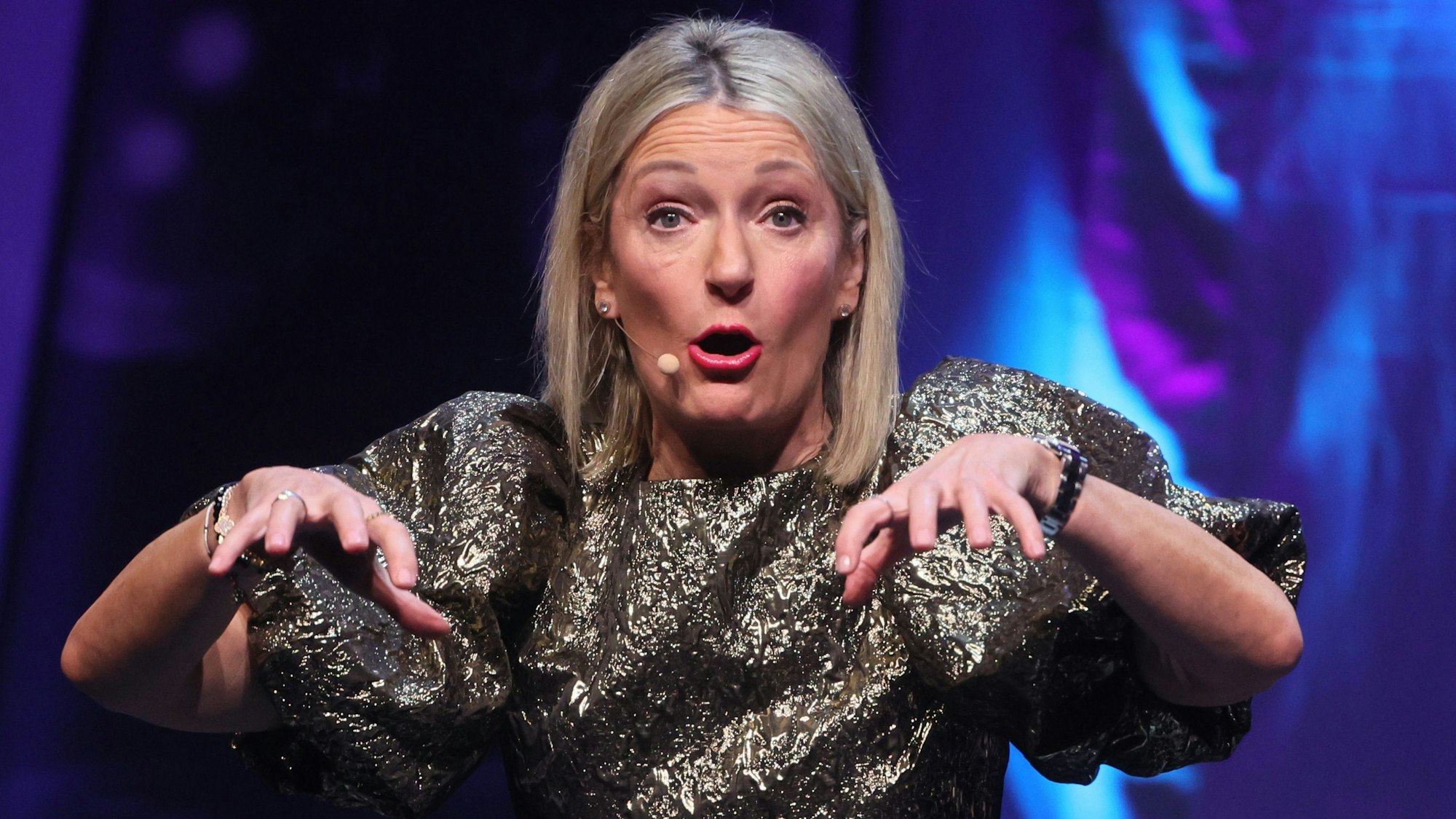 Die Kabarettistin Monika Gruber steht mit hocherhobenen Händen und geöffnetem Mund auf der Bühne. Sie trägt einen golddurchwirkten Anzug.