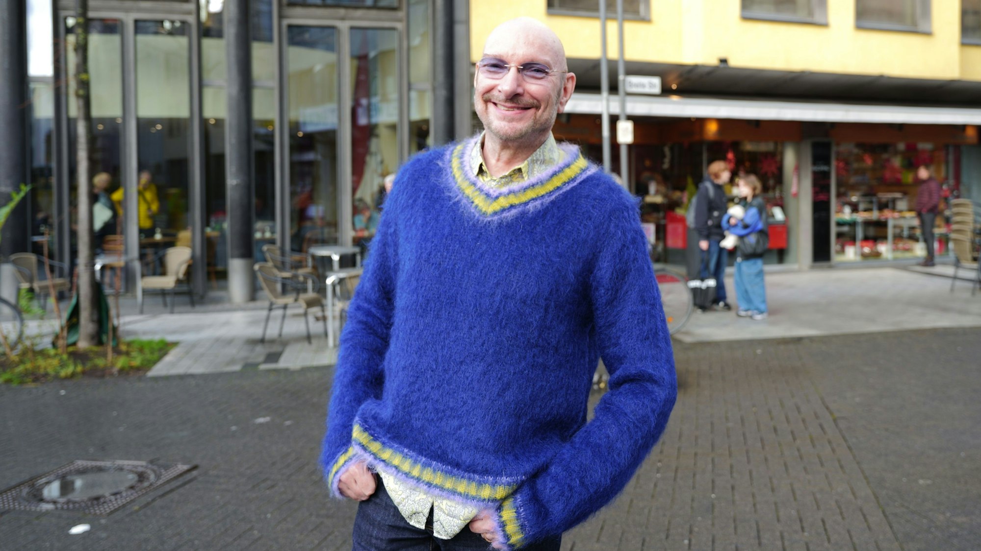 Schauspieler Ralph Morgenstern mit Glatze, leicht getönter Brille und knallig blauem Pullover auf der Breitestraße.