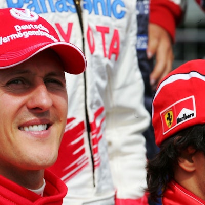 Michael Schumacher lächelt. Er trägt seinen roten Rennanzug und eine rote Kappe aus seiner Zeit bei Ferrari.