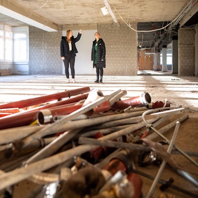 Das Bild zeigt zwei Schülerinnen in dem Rohbau. Kabel hängen von der Decke und Rohre sowie Leitungen liegen auf dem Boden.