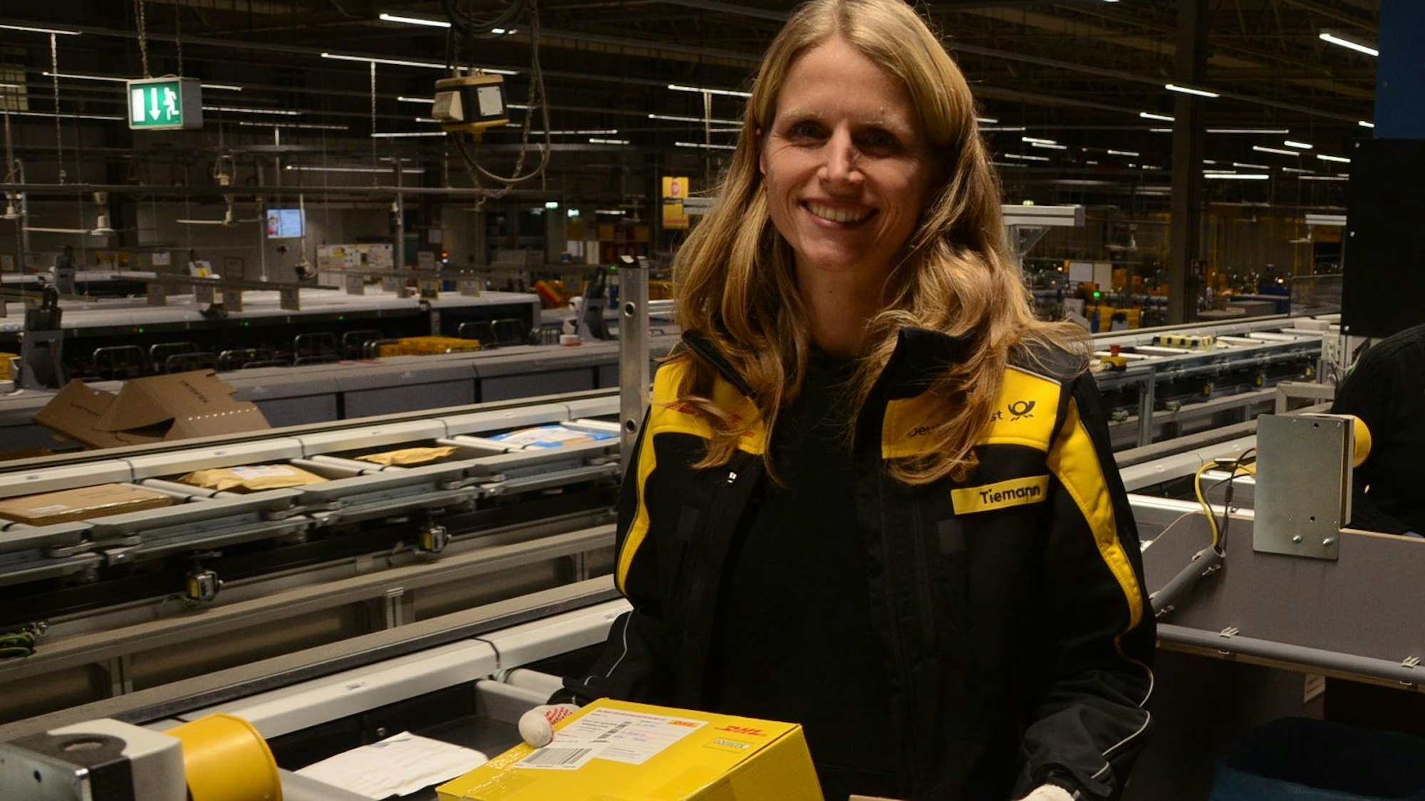 Eine Frau mit langen blonden Haaren sortiert Päckchen an einem Transportband.