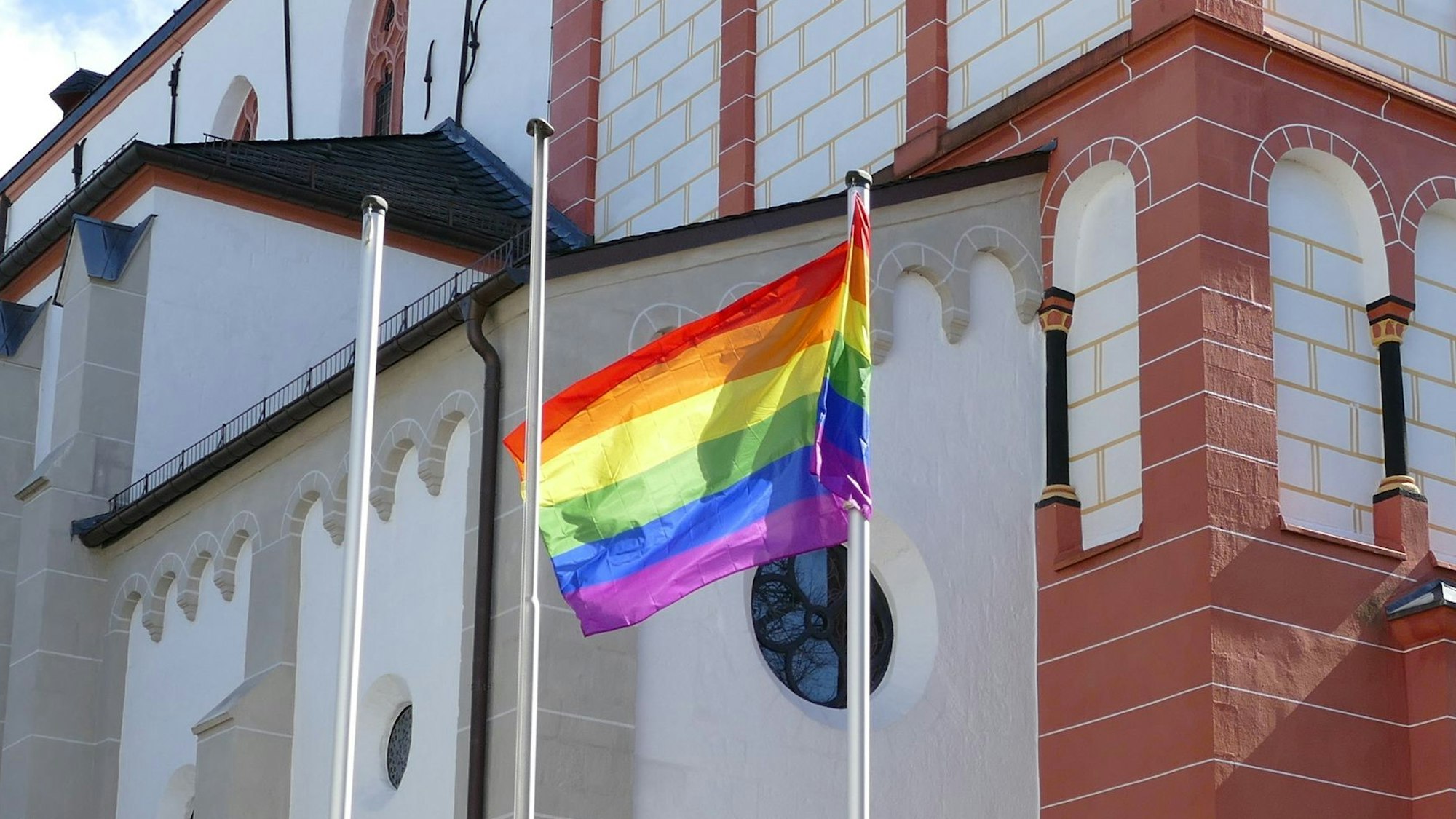 Eine Regenbogenflagge vor einer Kirche mit weiß-ziegelfarbener Fassade