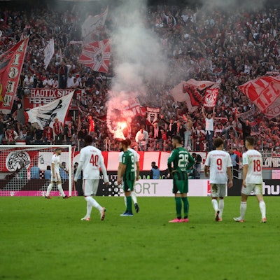 Während auf der Südtribüne des 1. FC Köln Bengalos brennen, wurde das Spiel gegen Borussia Mönchengladbach wegen Sichtbeeinträchtigung durch den erzeugten Rauch unterbrochen. Spieler beider Mannschaften stehen auf dem Rasen und betrachten das Treiben der Fans.