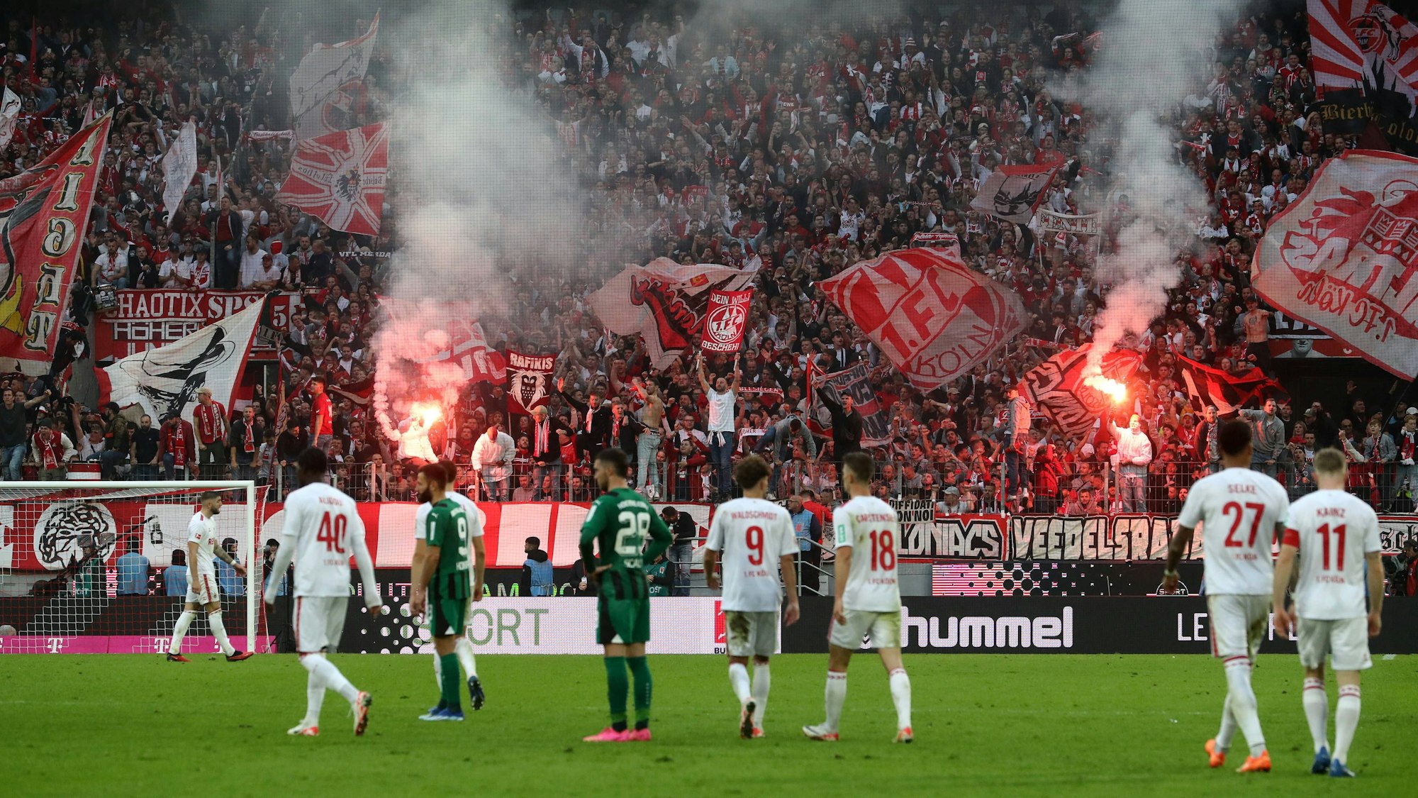 Während auf der Südtribüne des 1. FC Köln Bengalos brennen, wurde das Spiel gegen Borussia Mönchengladbach wegen Sichtbeeinträchtigung durch den erzeugten Rauch unterbrochen. Spieler beider Mannschaften stehen auf dem Rasen und betrachten das Treiben der Fans.