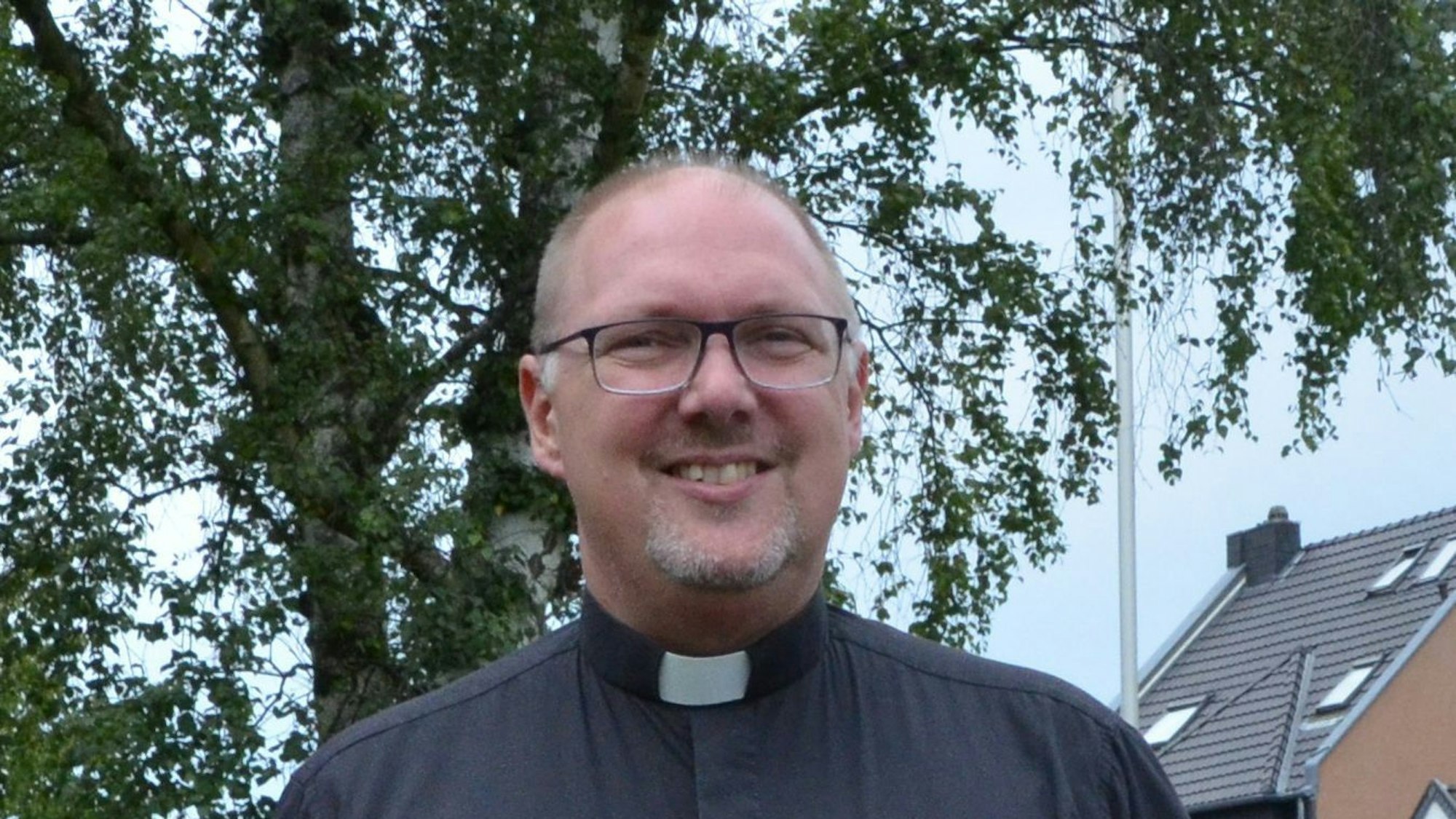 Ein Pfarrer mit schwarzem Hemd und weißem Kragenband vor einem Baum
