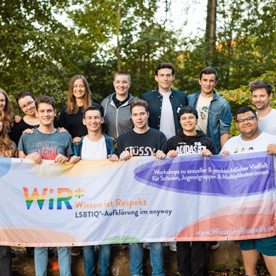 13 jugendliche Multiplikatorinnen und Multiplikatoren des „WiR*"-Projekts, das das „anyway “Kölner Jugendlichen anbietet, halten eine Fahne hoch, auf der „Wissen ist Respekt“ geschrieben steht.