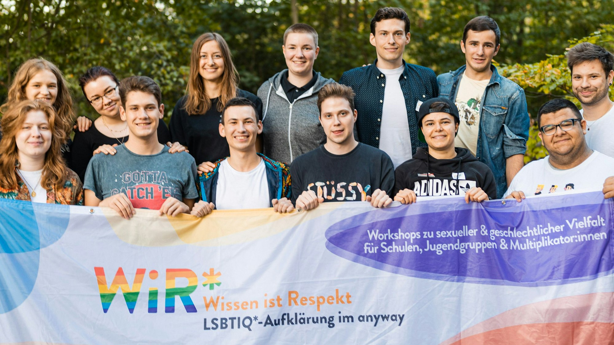 13 jugendliche Multiplikatorinnen und Multiplikatoren des „WiR*"-Projekts, das das „anyway “Kölner Jugendlichen anbietet, halten eine Fahne hoch, auf der „Wissen ist Respekt“ geschrieben steht.