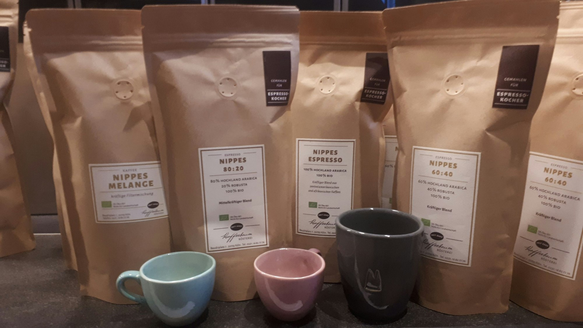 Kaffee-Kreationen in braunen Tüten in der Rösterei „Kaffeebaum“. Davor stehen Keramiktassen.