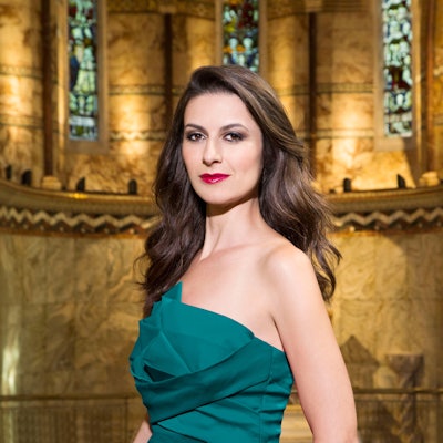 Chen Reiss trägt ein grünes Kleid und hat lange, gewellte braune Haare. Hinter ihr eine marmorne Wand eines alten Gebäudes.