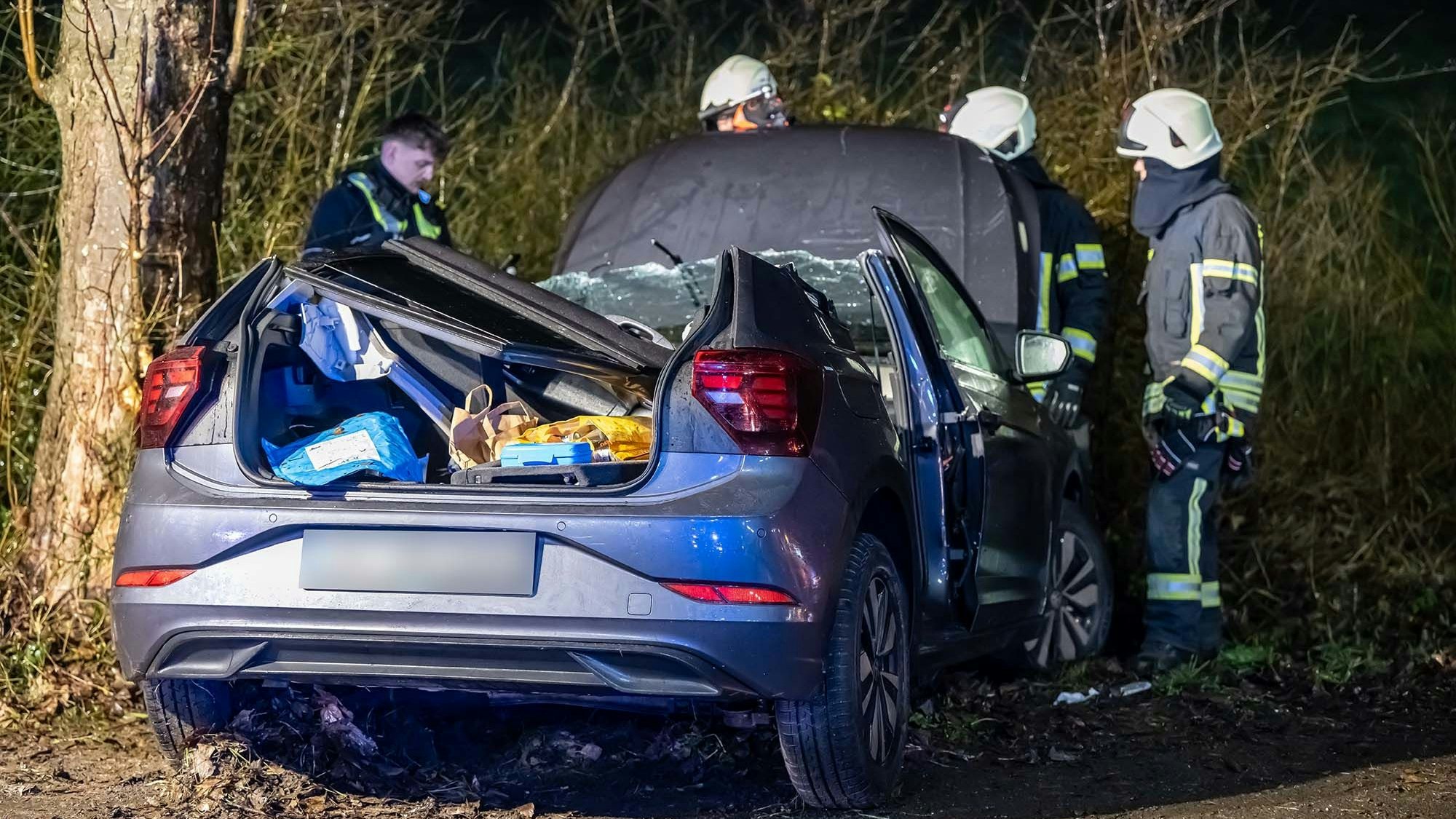 Ein Unfallwagen nach einem Aufprall gegen einen Baum. Mehrere Feuerwehrleute stehen um den Wagen herum.