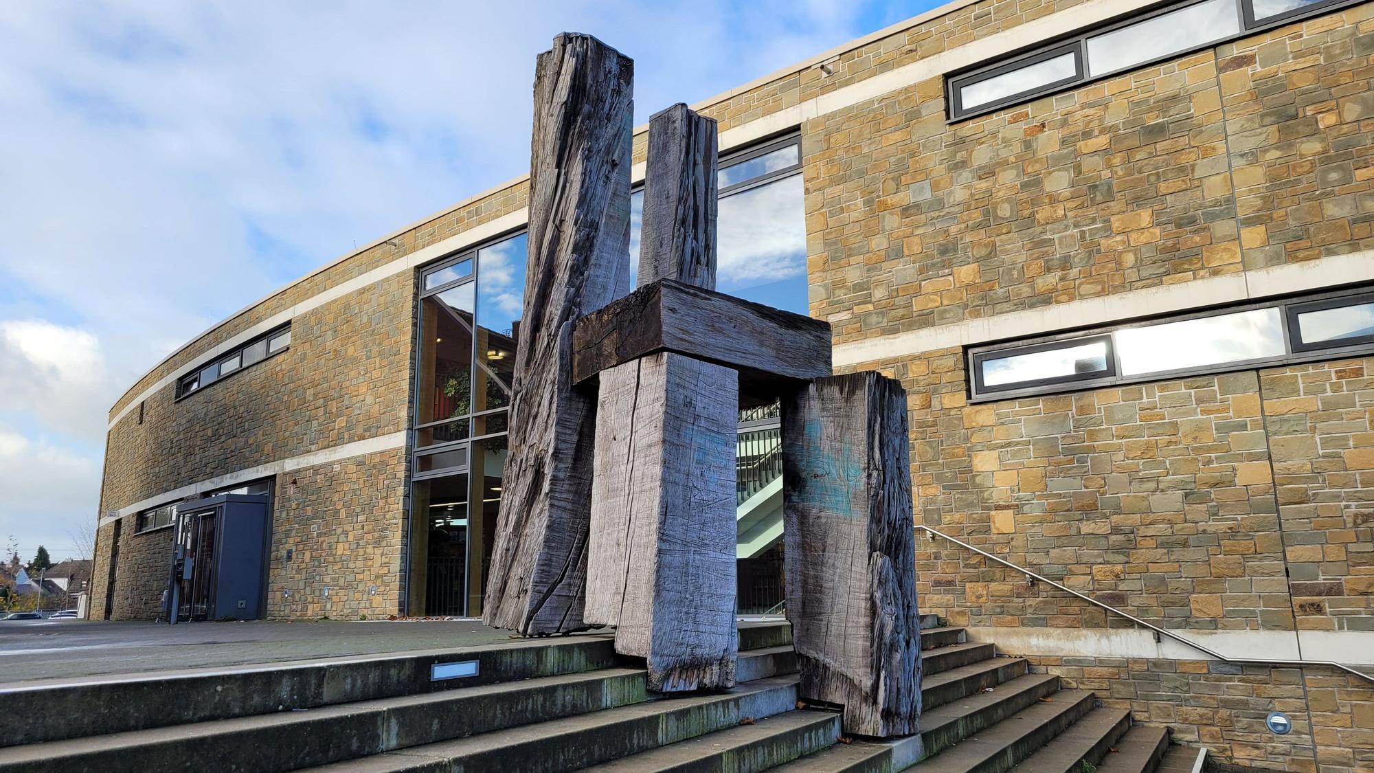 Zu sehen ist ein riesiger Stuhl aus Holz, er steht auf einer Treppe, im Hintergrund ein Gebäude mit einer hellen Klinkerfassade und Fenstern.