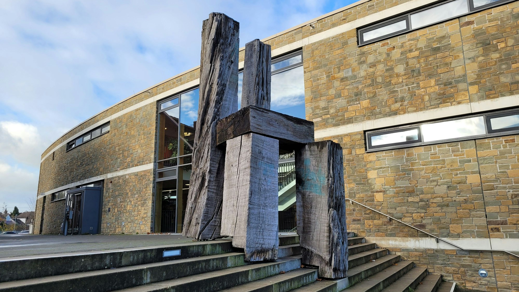Zu sehen ist ein riesiger Stuhl aus Holz, er steht auf einer Treppe, im Hintergrund ein Gebäude mit einer hellen Klinkerfassade und Fenstern.