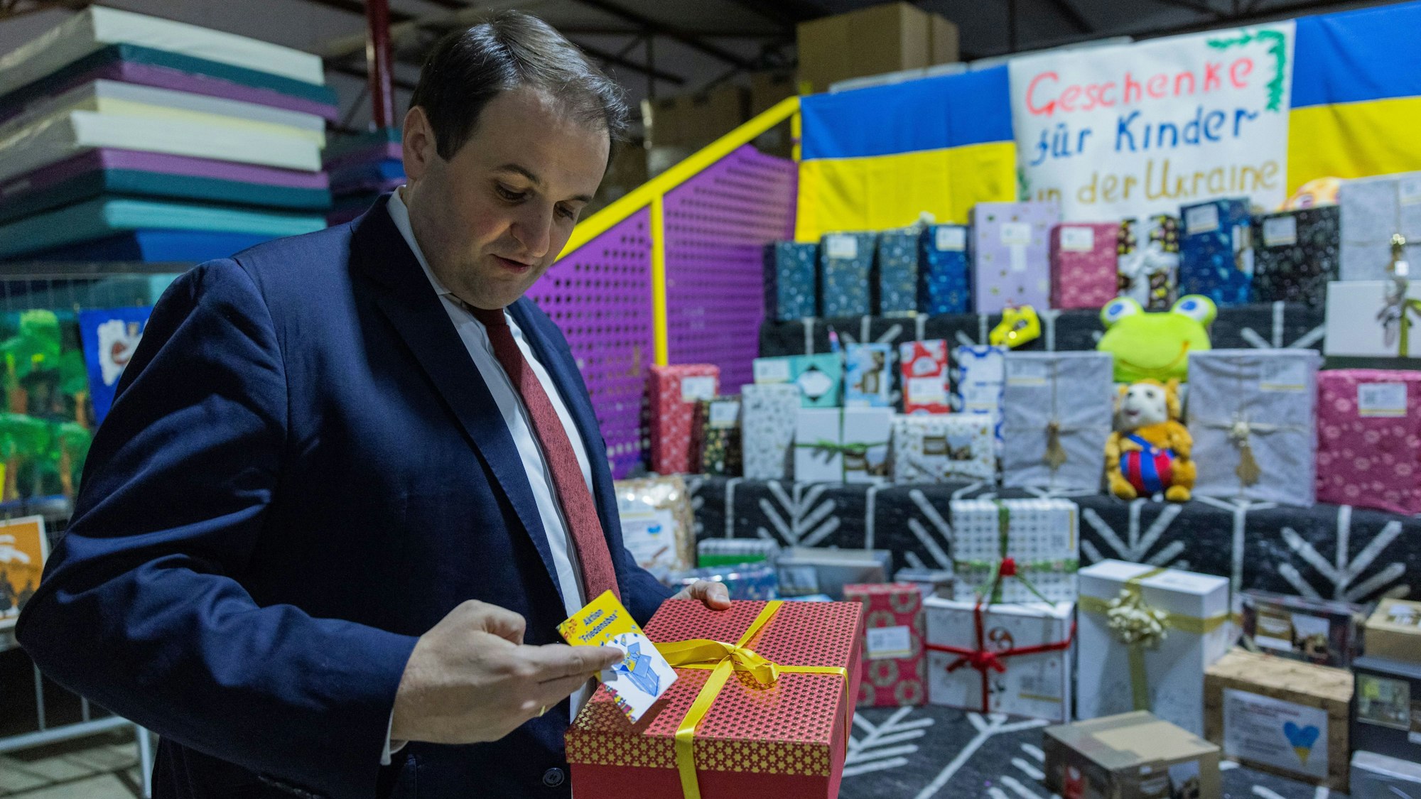 Mann mittleren Alters in dunkelblauem Anzug, hellblauem Hend und roter Krawatte, der ein Geschenk in der Hand hält und vor einem Berg mit Geschenken steht.