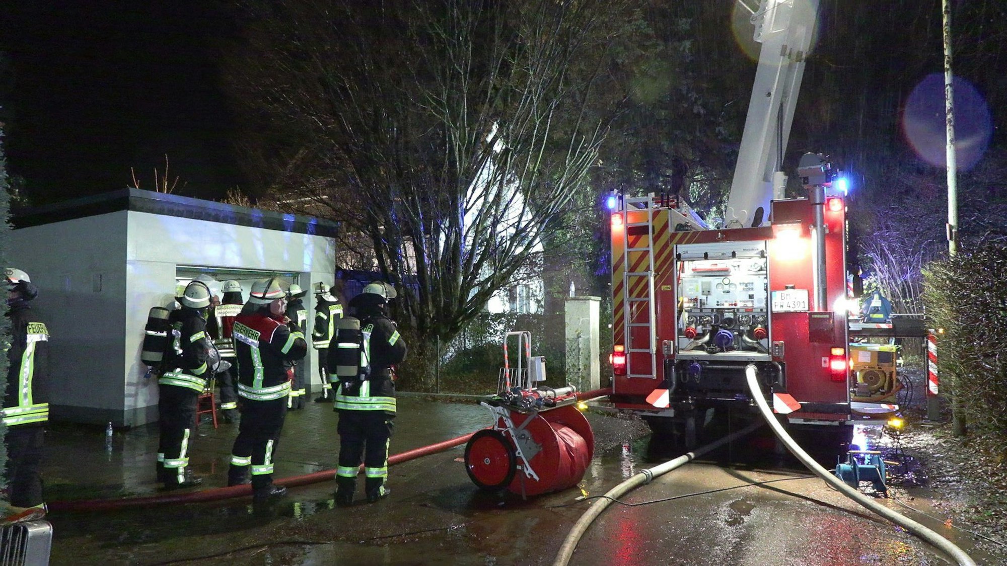 Ein Feuerwehreinsatzwagen steht vor einem Einfamilienhaus in Elsdorf. Einsatzkräfte stehen daneben. Schläuche ragen aus dem Wagen heraus, Scheinwerfer läuchten die Umgebung aus.