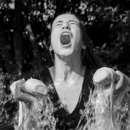 Schwarz-Weiß Fotografie einer jungen Frau, die Ballettschuhe in der hand hält. Sie wird mit Wasser übergossen und hat ihren Mund weit aufgerissen.