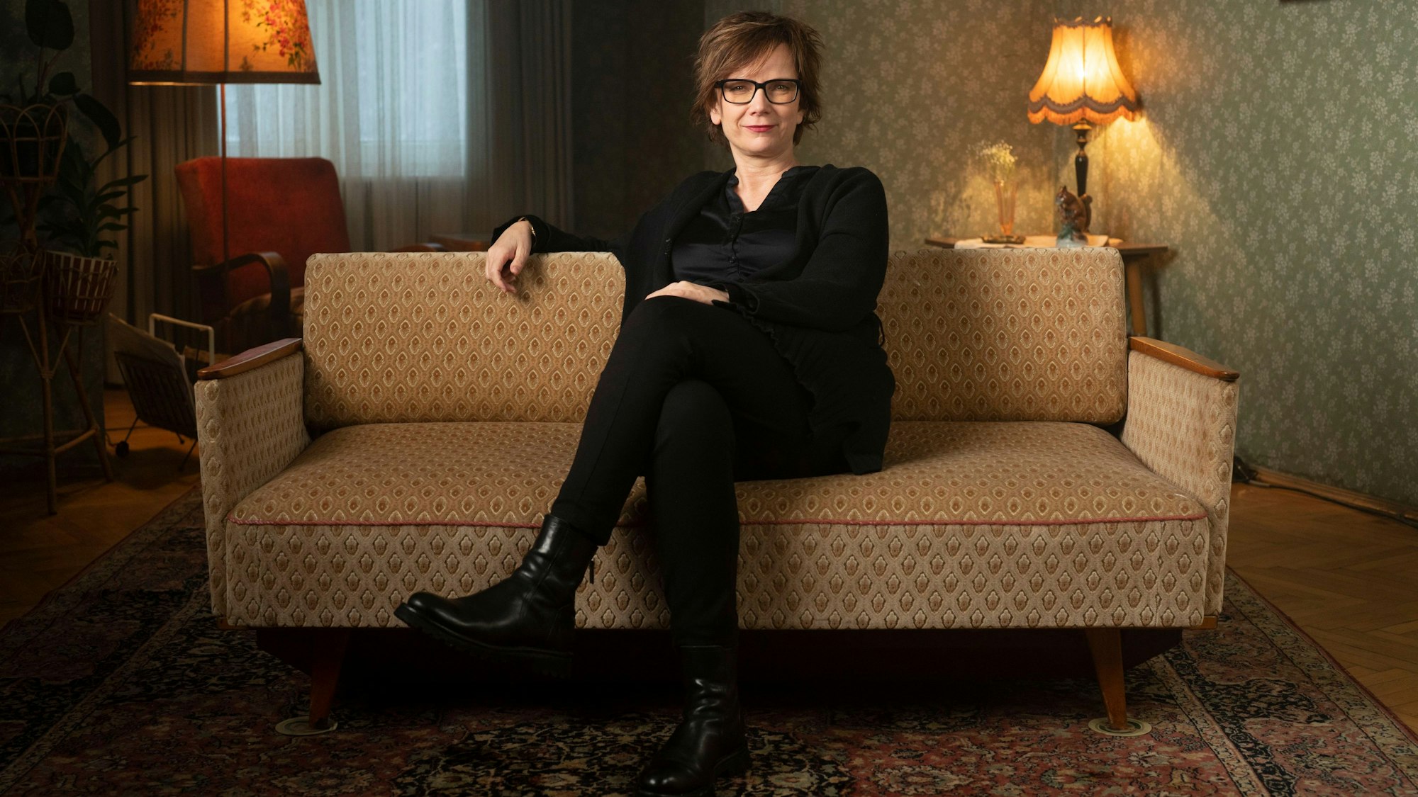 Sabine de Mardt sitzt in der Kulisse der Serie „Deutsches Haus“ auf einem Sofa, sie trägt schwarze Kleidung und eine schwarze Brille.